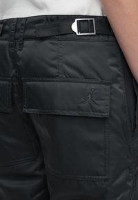 Close-up van zwarte broek met een klepzak, voorzien van een gestikt logo van een basketbalspeler en een verstelbare riem met een metalen gesp.