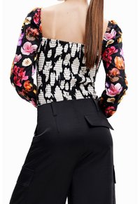 Schwarzes Oberteil mit floralem Muster und Ärmeln, kombiniert mit hoch taillierten schwarzen Hosen. Strukturierter Stoff, quadratischer Ausschnitt und Rückendetails sichtbar.