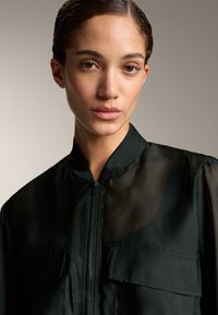 Musta vetoketjullinen takki, jossa on kaulus ja kaksi rintataskua; kevyt, puoliläpinäkyvä kangas, jossa on sileä pinta.