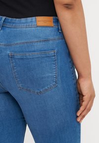 Blue denim jeans with a smooth texture, featuring a brown leather tag and a back pocket, tailored fit with subtle stitching details.