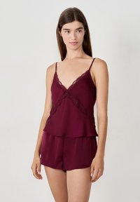 Ung kvinna klädd i burgunderfärgad camisole med spetskant och matchande shorts, stående mot en enkel ljus bakgrund, tittande något åt höger.