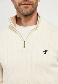 Cremfarbener Pullover mit Kabelstrickmuster und einem halben Reißverschluss am Kragen. Merkmale sind das strukturierte Muster und ein kleines, dunkelblaues gesticktes Logo auf der linken Brust.