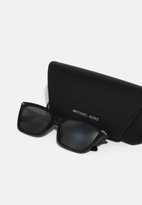 Juodi Michael Kors saulės akiniai su kvadratiniais lęšiais, matinio paviršiaus ir sidabriniais akcentais ant kojelių, kartu su juodu dėklu.