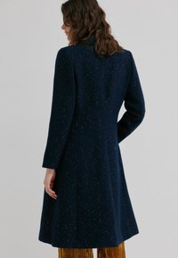 Femme aux cheveux bruns ondulés portant un long manteau bleu foncé à motifs tachetés et un pantalon couleur moutarde, vue de dos sur un fond clair uni.