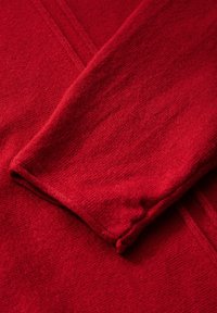 Röd, texturerad tyg med en mjuk stickad yta. Har en vikt ärmskant och ett subtilt mönster av horisontella linjer i materialet.