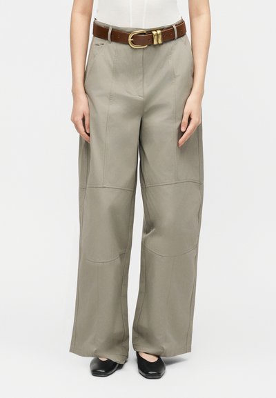 Femme portant un pantalon kaki à jambes larges avec des coutures à l'avant, une ceinture en cuir marron avec une boucle dorée, et des chaussures noires à bout carré.