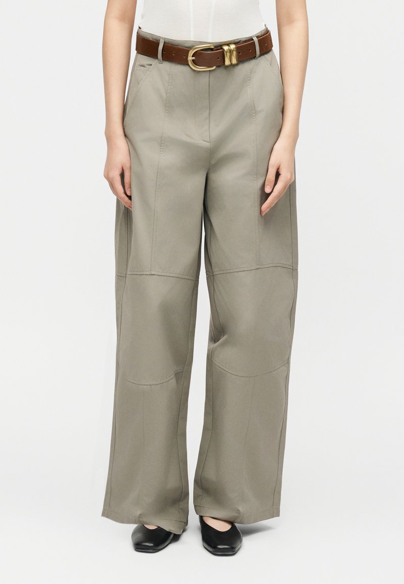 Donna che indossa pantaloni khaki a gamba larga con cuciture frontali, una cintura di pelle marrone con fibbia dorata e scarpe nere con punta quadrata.