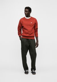 Sweatshirt vermelha com um logótipo Nike branco, combinada com calças cargo verde-oliva largas e ténis pretos, exibindo um visual casual.