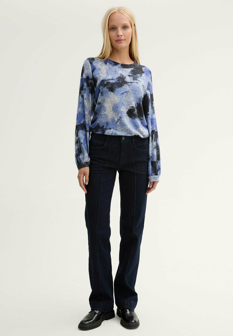 Langärmliges Hemd in Blau-Schwarz-Muster, kombiniert mit dunklen Jeans im Schlag. Das Hemd hat eine lockere Passform; die Jeans verfügen über Fronttaschen.