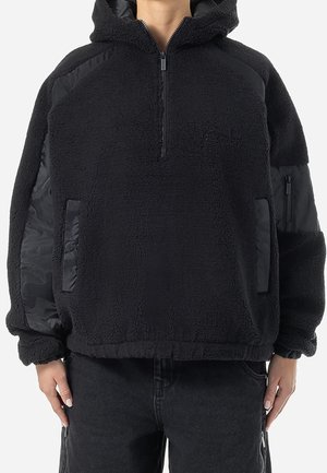 Czarna polarowa kurtka z kapturem, z półzamkiem, z podszytymi łatkami na ramionach i rękawach, przednimi kieszeniami i elastycznymi ściągaczami, noszona przez stojącą osobę.