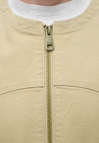 Veste beige à fermeture éclair avec tirette en laiton gravée "HUGO", portée sur une chemise blanche côtelée avec texte embossé "Porsche BOSS".