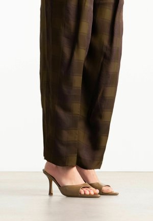 Personne portant un pantalon large à carreaux marron et des mules à talons hauts beiges à bout ouvert, debout sur un sol lisse.