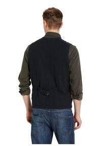 Granatowa bezrękawnik z bawełny, z detalem w postaci ściągniętego tylu, zestawiony z ciemnozieloną koszulą z długim rękawem oraz dżinsami.