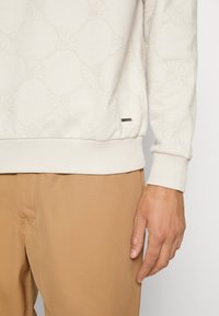 Sweat-shirt couleur crème avec un motif floral subtil, des poignets et un ourlet côtelés, ainsi qu'une petite étiquette métallique de la marque en bas.