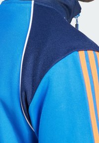 Sininen ja laivastonsininen verkkatakki oransseilla raidoilla, jossa on korkea kaulus, teksturoitu kangas ja kontrastiväriset paneelit. Vetoketjutasku kauluksessa.