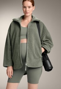 Zeleno-olivna flis jopica na zadrgo z teksturirano površino, nosena čez ujemajočo se športno modrček in kratke hlače, z črno jogamatko v roki.