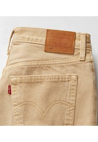 Jeans de mezclilla beige con una etiqueta de cuero, que presentan un logo, costura doble, un bolsillo trasero y una pequeña etiqueta roja en el lateral.