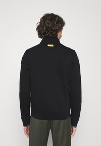 Svart tröja med hög krage, strukturerad tyg och en gul accent på ryggen. Funktioner inkluderar ribbade muddarna och en åtsittande passform.