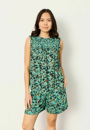 Młoda kobieta w turkusowej, kwiecistej bluzce bez rękawów i dopasowanych szortach stoi na jasnym, jednolitym tle z jedną ręką w kieszeni.