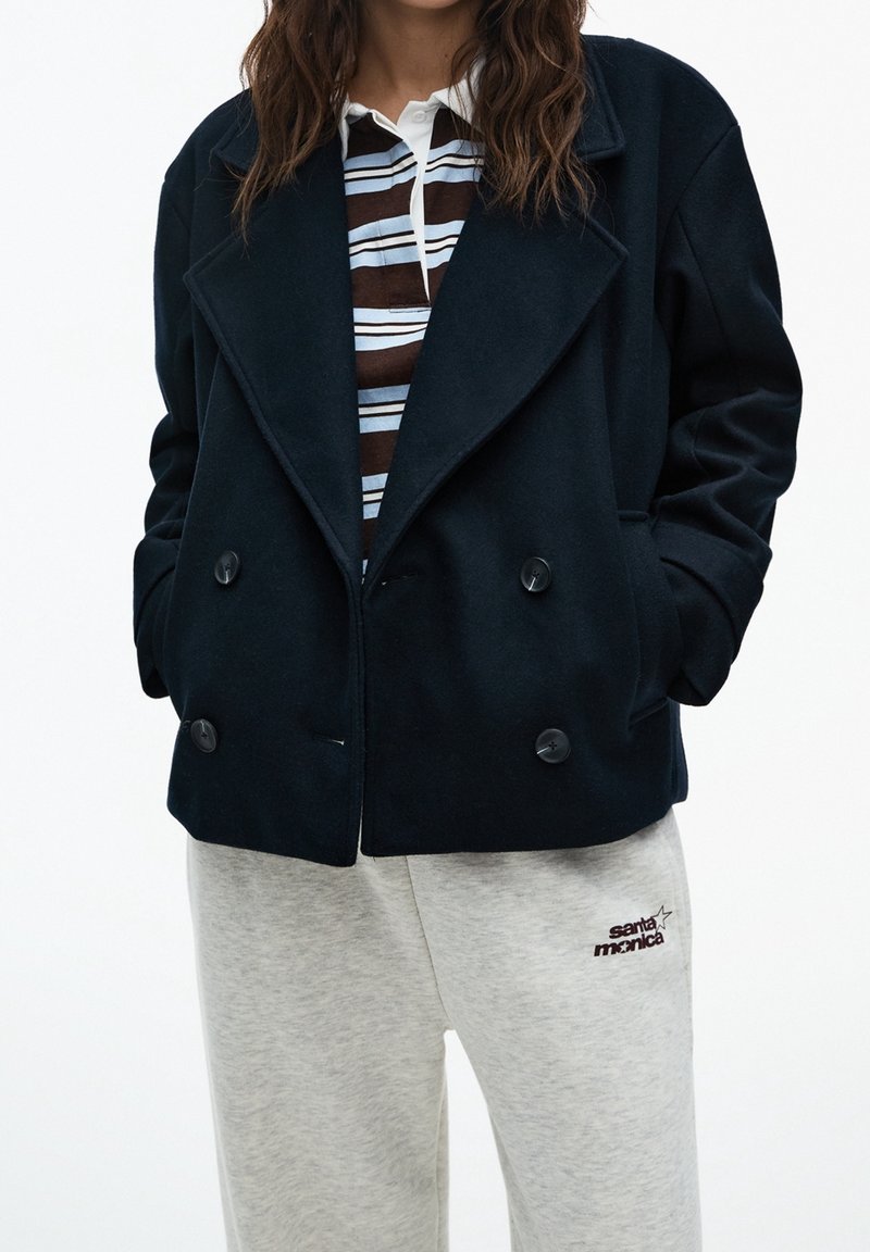 Granatowy płaszcz podwójnie zapinany z miękkiego materiału z dużymi klapami, w połączeniu z paskowanym polo i szarymi spodniami dresowymi z logo "santa monica".