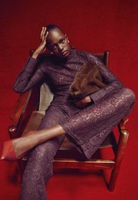 Purple lace jumpsuit with flared legs, paired with a brown faux fur clutch. Model seated on a wooden chair against a red background.