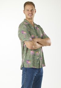 Camicia a maniche corte, con bottoni, di colore verde oliva con motivi di palme multicolori. Materiale in cotone con vestibilità rilassata, dotata di colletto da campeggio.