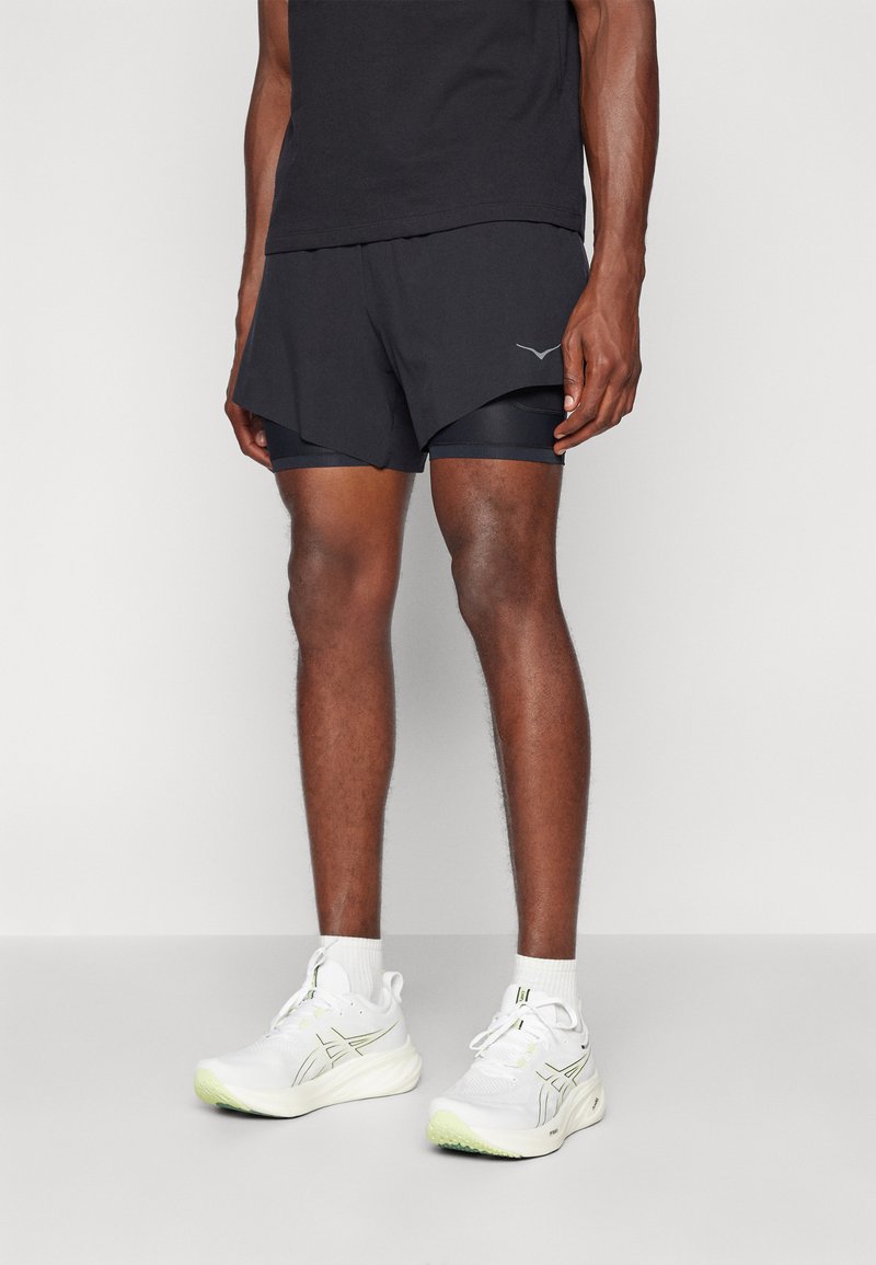 Șorturi atletice negre cu căptușeală interioară, cu un tiv curbat și un logo. Adidași albi cu accente verzi și tălpi texturate.