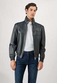 Άνδρας φορώντας ένα σκούρο γκρι δερμάτινο bomber μπουφάν πάνω από ένα λευκό T-shirt και σκούρα μπλε τζιν, στηρίζεται σε έναν απλό ανοιχτό φόντο.