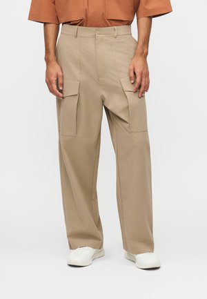 Άνδρας που φοράει μπεζ cargo παντελόνι με φαρδιά μπατζάκια, λευκά αθλητικά παπούτσια και καφέ κοντομάνικο πουκάμισο, στέκεται μπροστά σε λευκό φόντο.