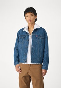 Chaqueta de mezclilla azul con un patrón de paisley, con cierres de botón, bolsillos laterales y un cuello clásico, llevada sobre una camisa blanca y pantalones marrones.