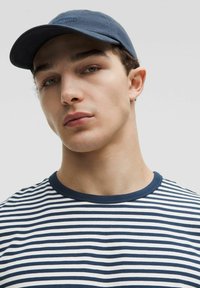 Casquette marine avec logo, t-shirt rayé blanc et marine, design à col ras du cou, texture lisse, vue en gros plan de la partie supérieure du corps, fond neutre.