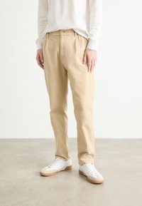 Beige, laza szabású pamutnadrág, rugalmas derékpánttal és két elülső zsebbel, fehér, gumi talpú sportcipővel párosítva.
