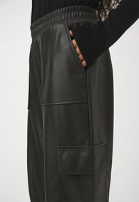 Osoba v černých kalhotách z umělé kůže s velkými náprsními kapsami, s jednou rukou vloženou v přední kapse a s černým dlouhým rukávem.