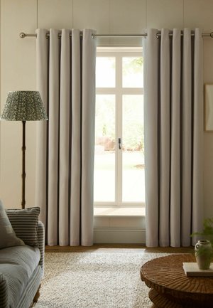 Hellbeige Vorhänge hängen an einem Fenster mit einer Holz-Stehlampe links und einer gestreiften Sofa-Ecke daneben.