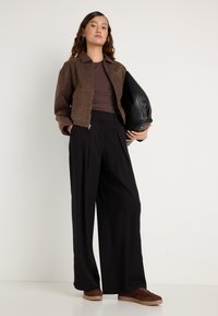 Casaco castanho com padrão sobre uma blusa castanha, combinado com calças pretas de cintura alta e sapatilhas castanhas, segurando uma mala preta.