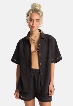 Camicia nera leggera con bottoni e maniche corte, rifinita con inserti traforati simili ad un pizzo, abbinata a pantaloncini con vita elastica. Stile casual da spiaggia.