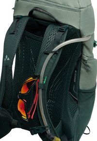 Sac à dos en tissu vert avec des sangles en maille, équipé d'un tuyau d'hydratation, de lunettes de soleil avec des accents rouges et d'une étiquette en matériaux recyclés.