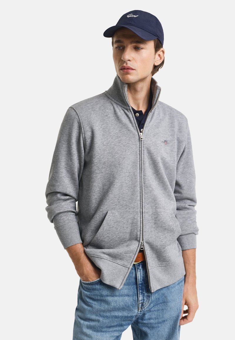 Sudadera gris con cremallera y alto cuello, con bolsillos laterales, puños acanalados y un logo en el pecho, llevada sobre una camiseta azul marino y unos jeans de mezclilla.