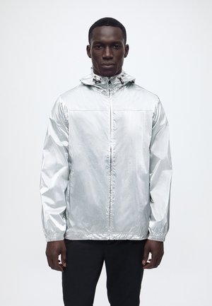 Man draagt een glanzend zilverkleurig capuchonjack dat dichtgeritst is, staat tegen een effen lichte achtergrond, met handen ontspannen langs het lichaam.