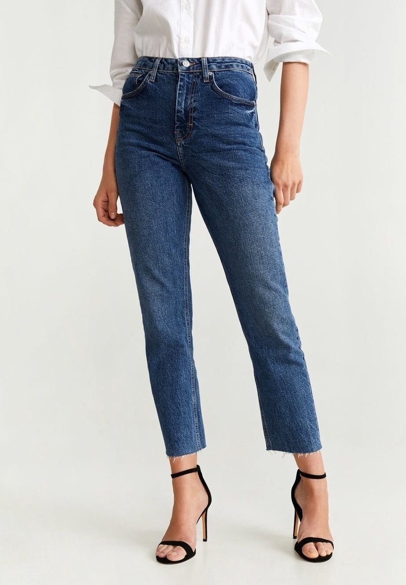 Femme portant un jean en denim bleu taille haute avec ourlet brut, associé à des sandales à talons hauts noires avec bride cheville et une chemise blanche à manches longues.