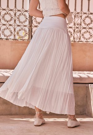 Kobieta w lekkiej białej plisowanej spódnicy i wiązanych płaskich butach, stojąca w pomieszczeniu obok ozdobnej balustrady z kutego żelaza.