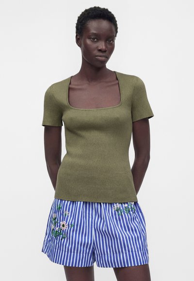 Γυναίκα φοράει ελαφρώς ριμπ πλεκτό τοπ ελιάς με κοντά μανίκια και τετράγωνο ντεκολτέ, καθώς και μπλε-άσπρα ριγέ σορτς με κεντημένα λουλούδια.