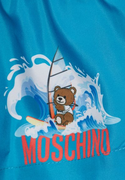 Bañador de color turquesa con un oso de dibujos animados sobre una tabla de surf, rodeado de olas. Texto "MOSCHINO" en rojo debajo de la imagen. Tela suave y ligera.
