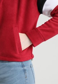Punainen fleece-pusero, jossa on mustavalkoisia yksityiskohtia, sivutasku ja mukava istuvuus. Näkyvät saumat ja teksturoitu kangas.