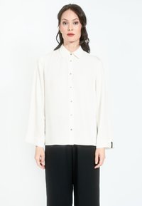 Biała bluzka z długim rękawem, z kołnierzykiem i zapinana na guziki, w połączeniu z czarnymi spodniami. Gładka faktura i subtelny połysk.