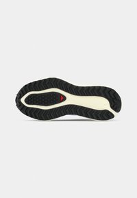 Semelle de chaussure de sport avec une semelle extérieure en caoutchouc noir présentant un motif de bande de roulement en zigzag, une semelle intermédiaire crème, et un accent rouge près du talon.