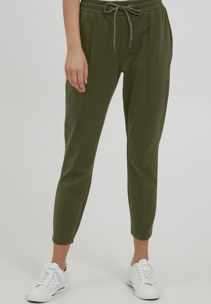 Maslinasto zelene tapered joggers hlače od mekog materijala, s elastičnim pojasom i vezicom, te bočnim džepovima, u kombinaciji s bijelim tenisicama.