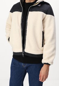 Fleece-takki, jossa on vaaleanbeige vartalo, mustat nailonpaidat, vetoketjullinen etuosa, joustavat hihansuut ja sivutaskut. Teksturoitu, lämmin ja rento muotoilu.