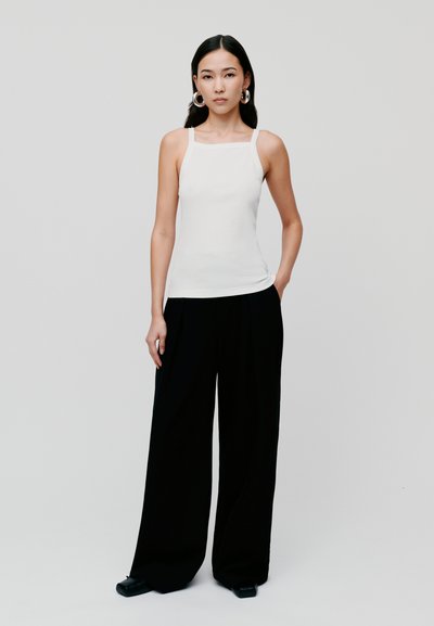 Top fără mâneci alb, ajustat, cu decolteu pătrat, asortat cu pantaloni largi negri. Modelul stă pe un fundal neutru, cu mâinile în buzunare.