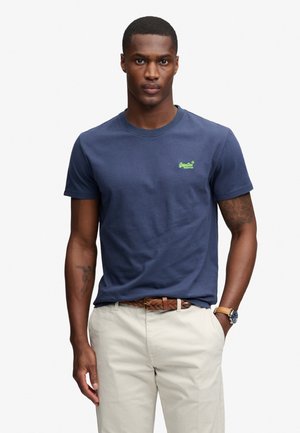Uomo che indossa una t-shirt blu navy a maniche corte con piccolo logo verde, pantaloni beige, cintura marrone intrecciata, orologio da polso, in piedi con una mano in tasca.